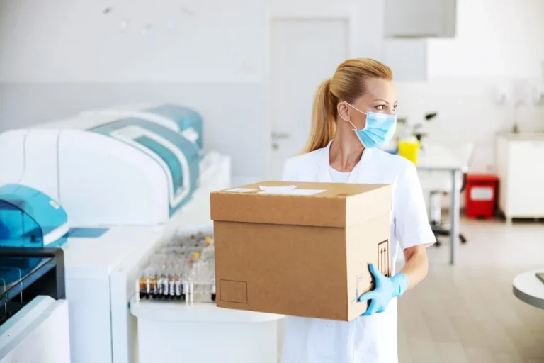 Laboratory technician in a lab coat and gloves carrying a sealed specimen transport box through a clinical laboratory